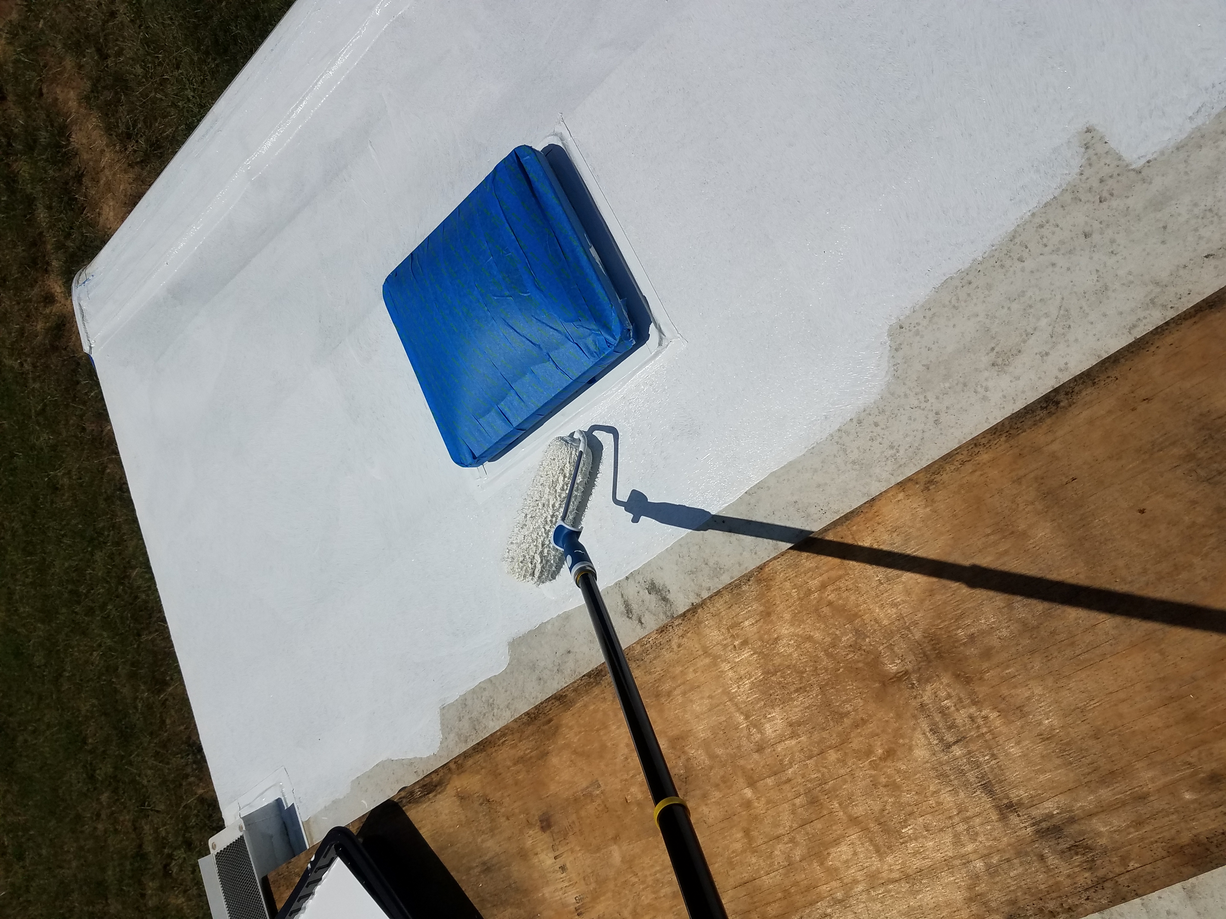 Painting the roof of the travel trailer with a long-handeled roller while standing on plywood