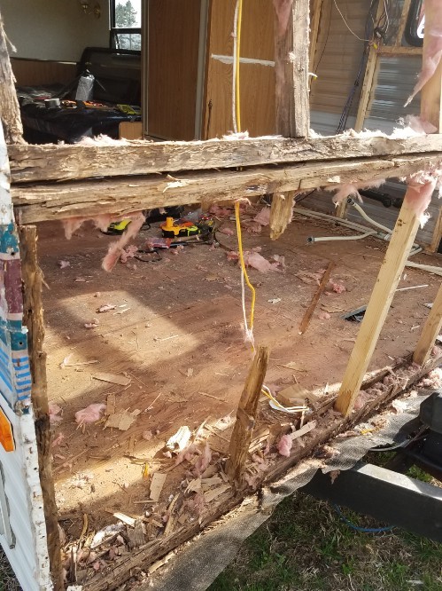 The frame of the travel trailer with the siding taken off, showing the wooden frame which is rotten and falling apart from water damage and some wiring.