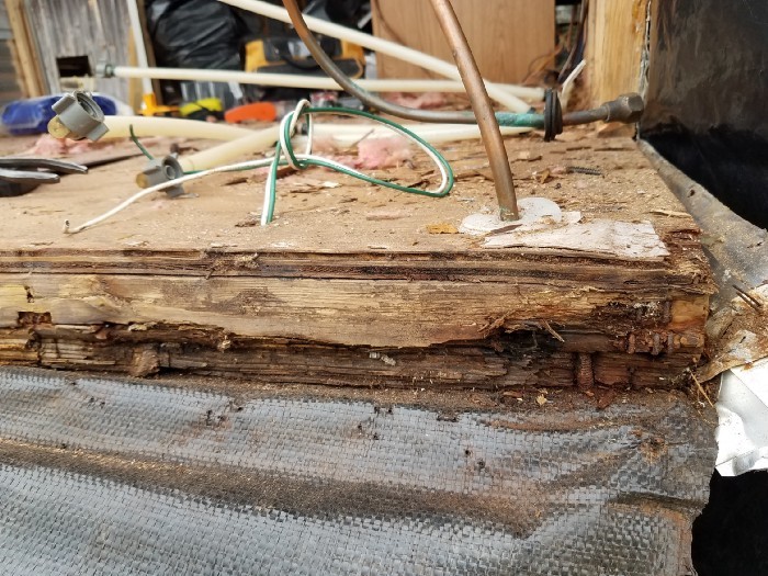 Rotted plywood floor with wires and copper pipes in the background.